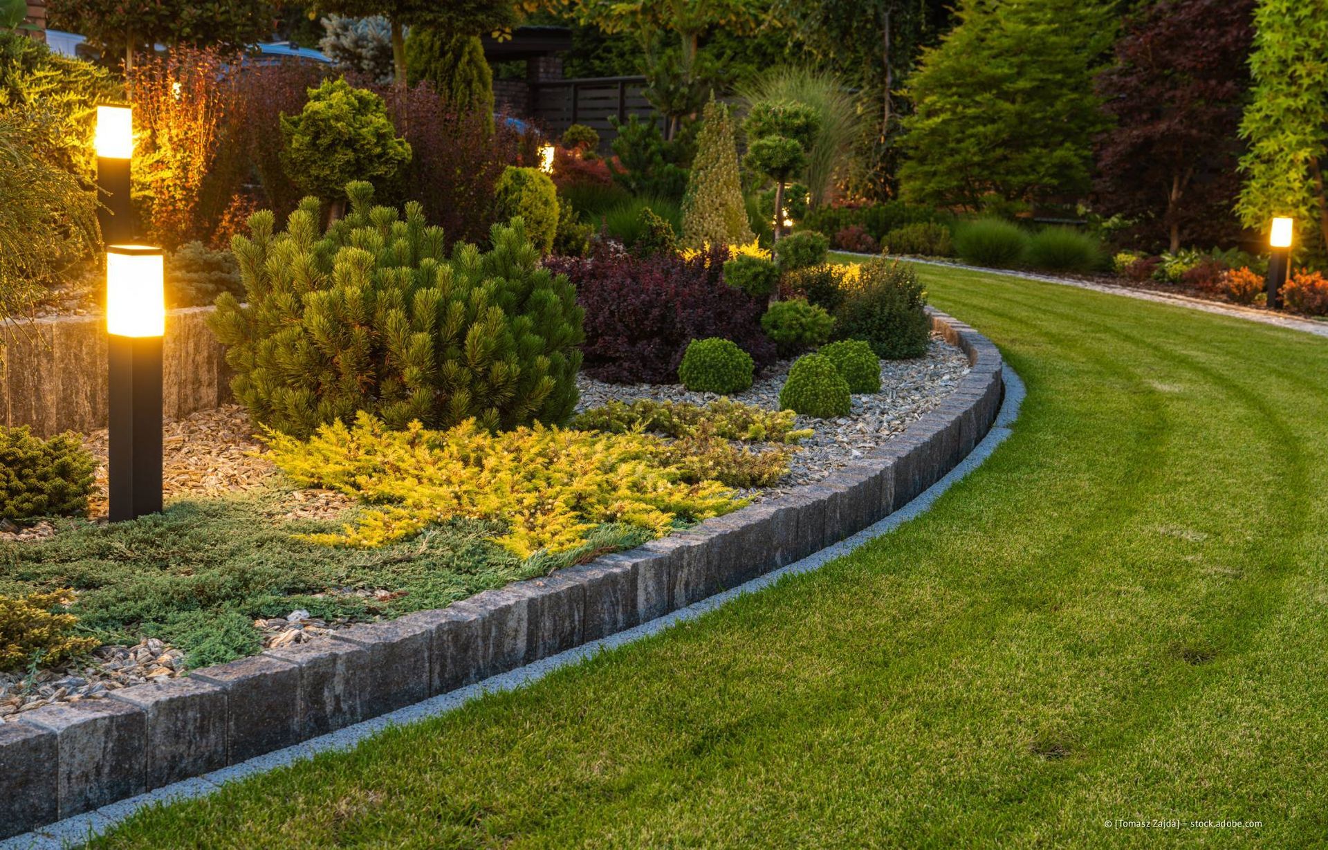 Üppiges Gartenbeet mit verschiedenen Pflanzen und Wegbeleuchtung entlang eines geschwungenen grünen Rasens.