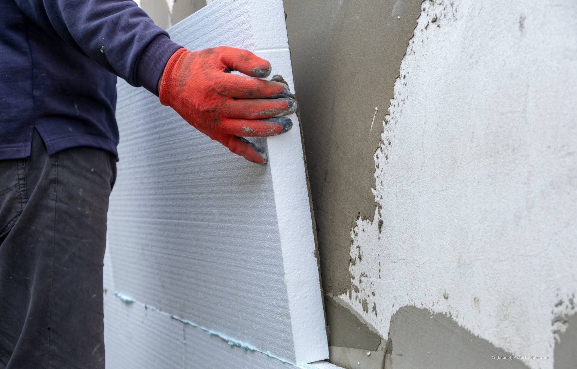 Eine Person mit roten Handschuhen montiert Dämmplatten mit aufgetragenem Klebstoff an einer Wand.