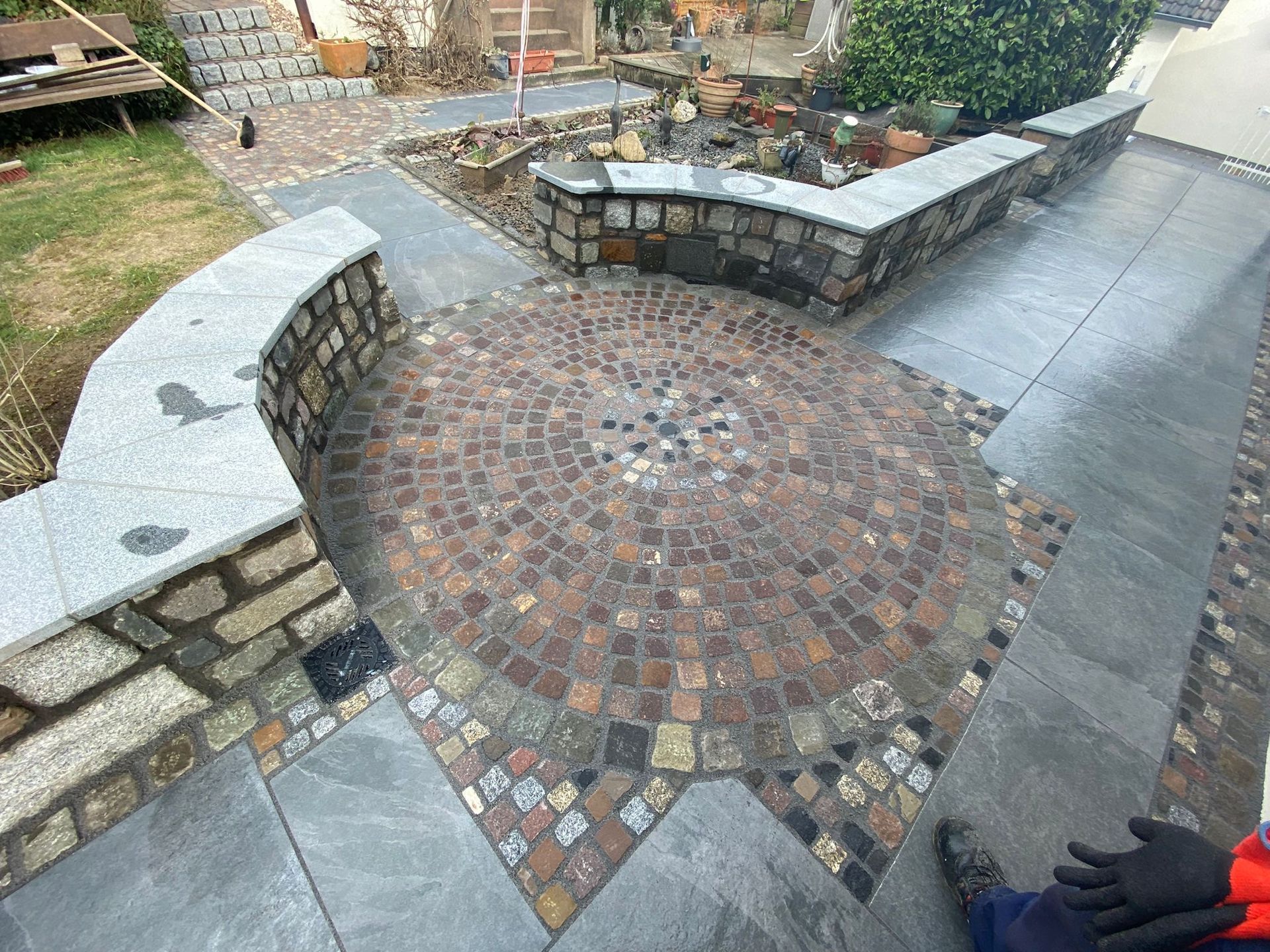 Runder, mit Kopfsteinpflaster belegter Patio mit Steinmauern und Stufen, die zu Gartenwegen führen.