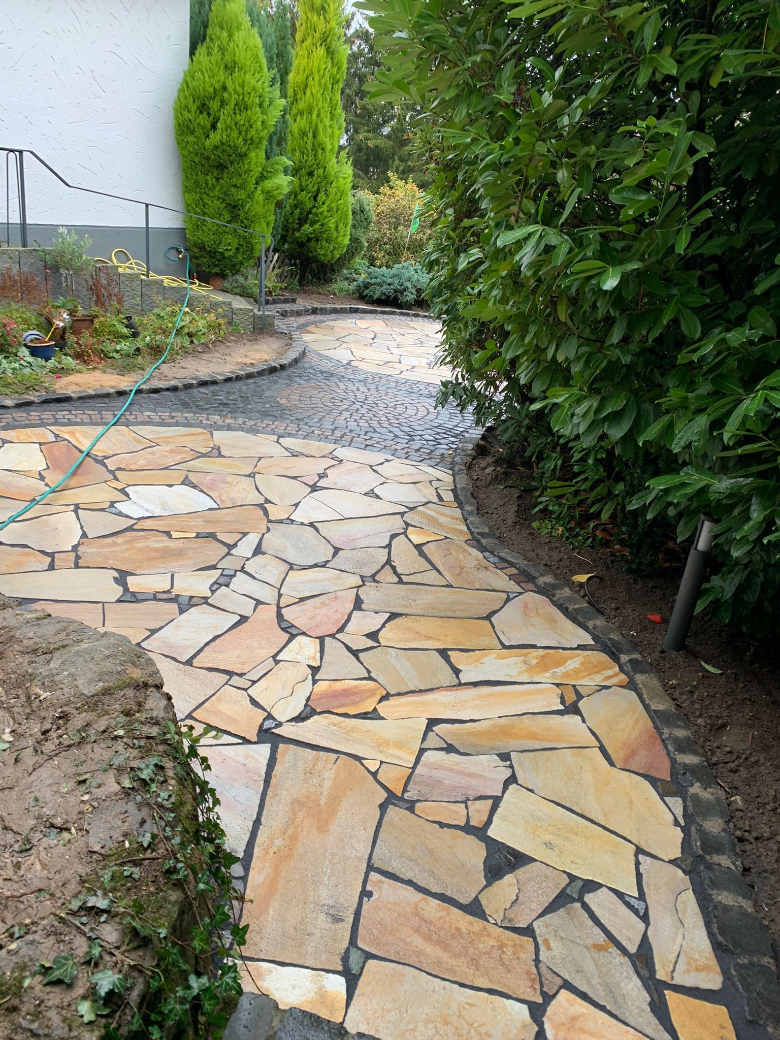 Ein mit Steinplatten belegter Weg führt durch einen Garten, der von Pflanzen und Bäumen gesäumt ist.