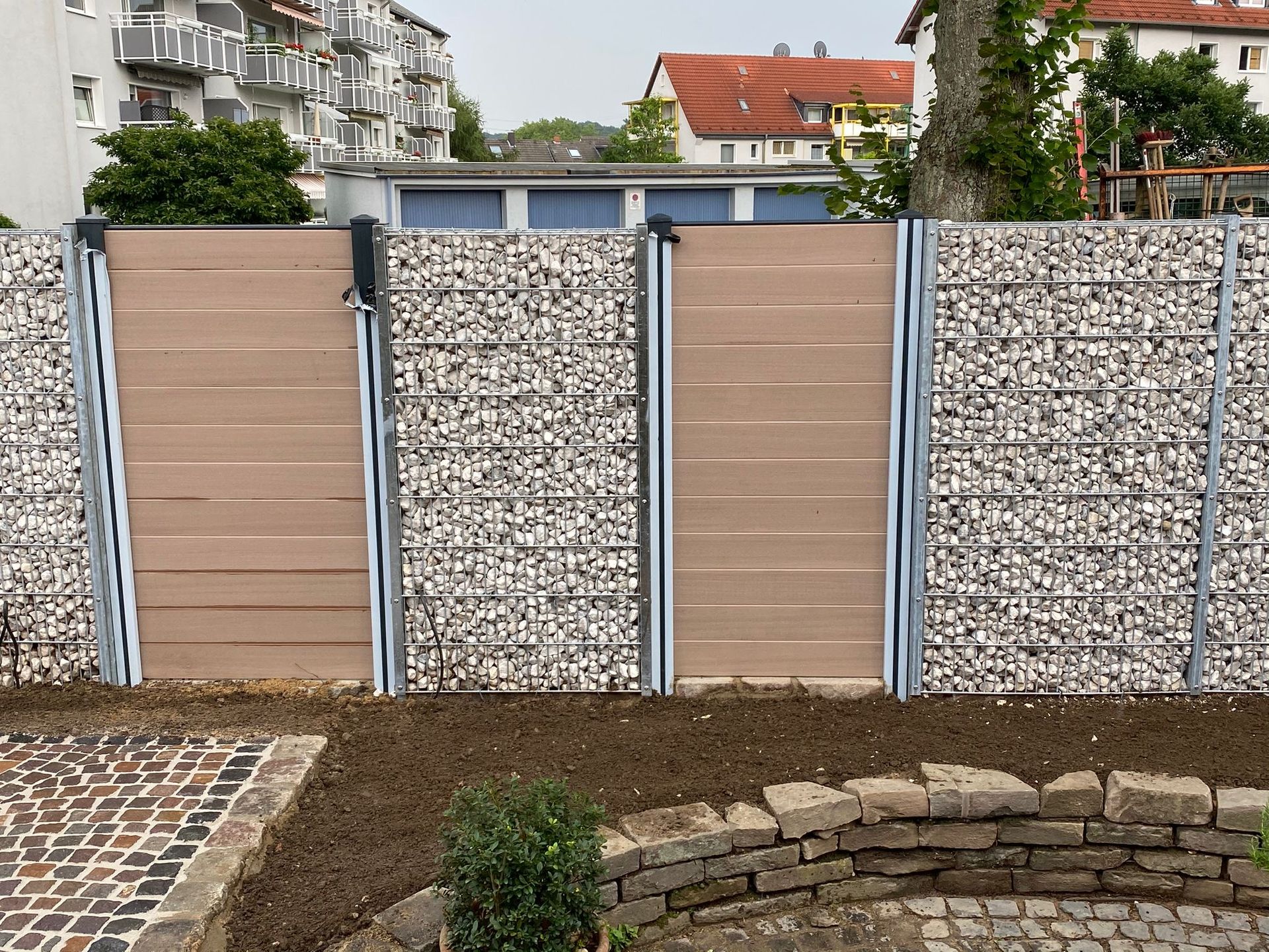 Ein provisorischer Zaun grenzt an eine Rasenfläche neben einem Schotterweg; im Hintergrund ist ein Gebäude zu sehen.