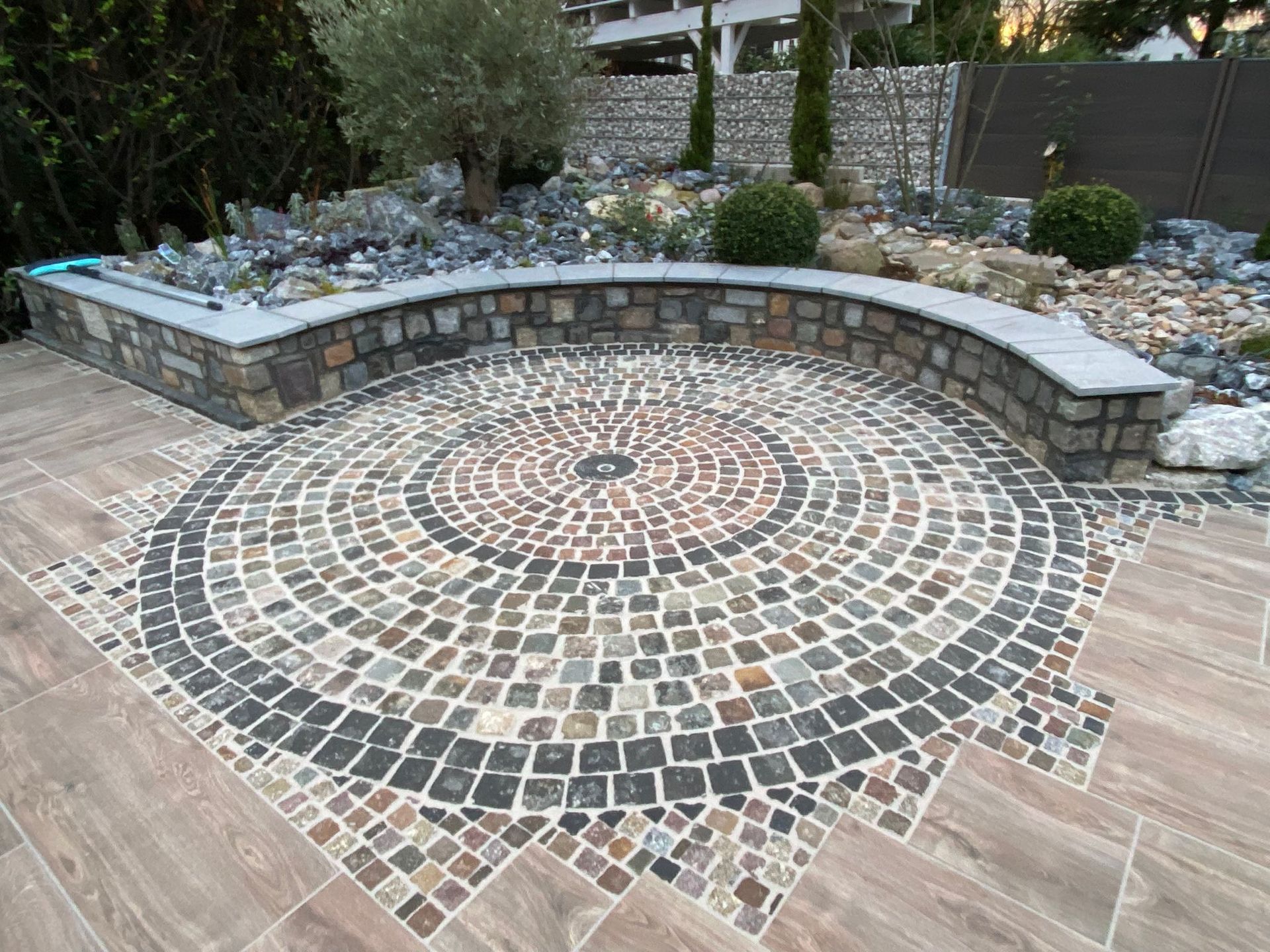 Runder, mit Kopfsteinpflaster belegter Patio mit Steinmauer und umliegender Landschaftsgestaltung.
