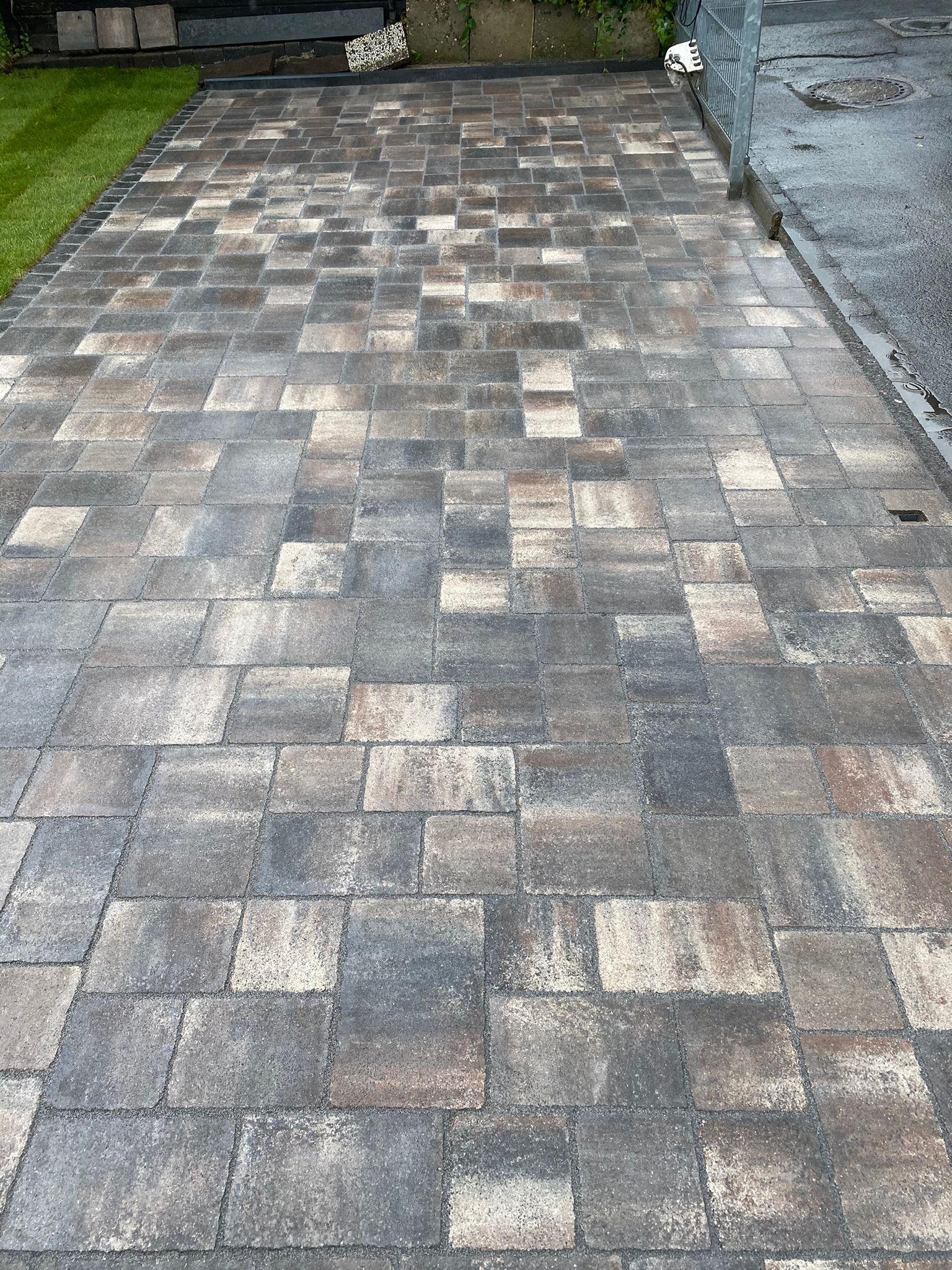 Ziegelpflaster in Grau- und Brauntönen bedeckt eine Einfahrt, die an einen grünen Rasenstreifen angrenzt.