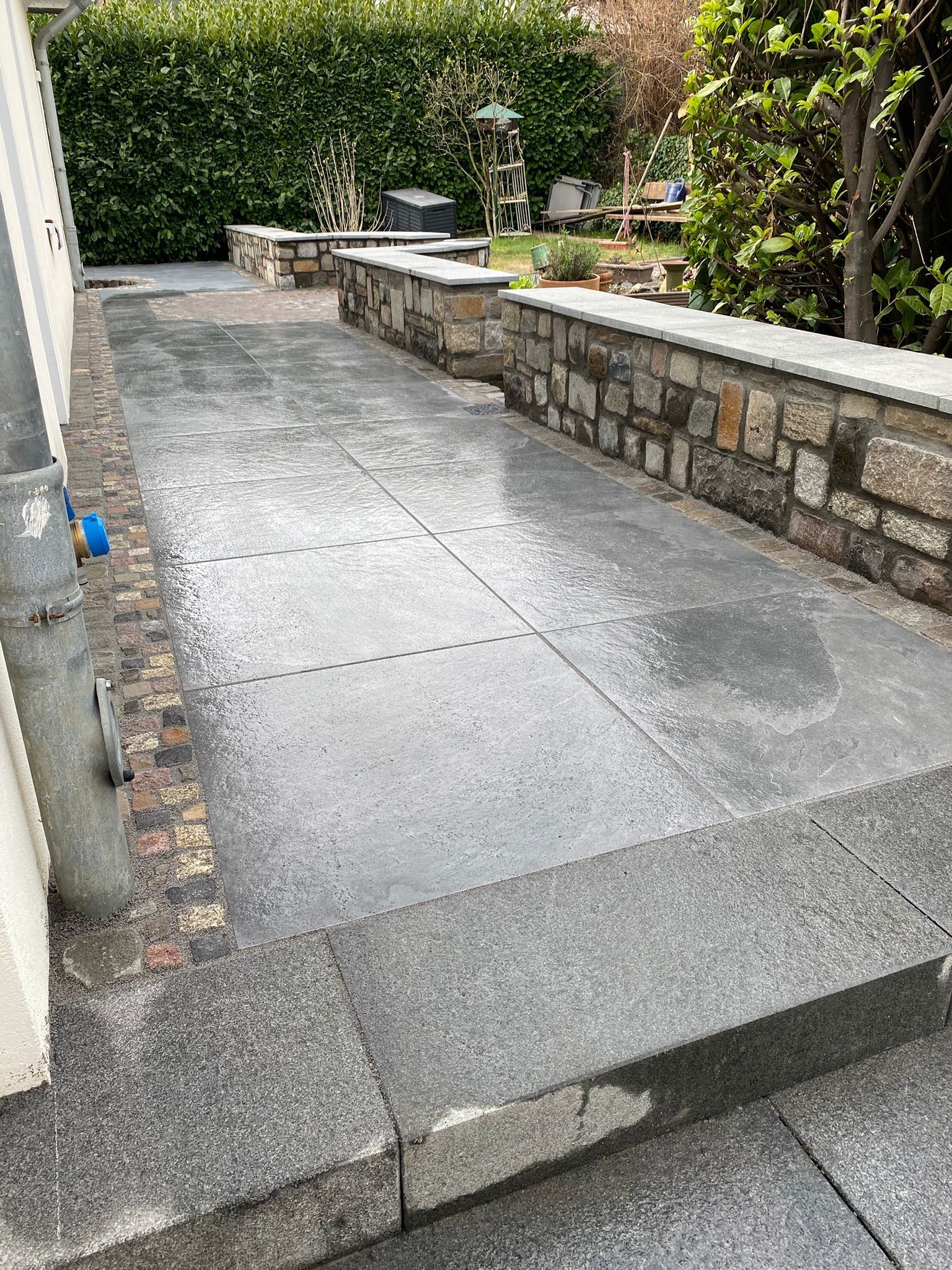 Steinterrasse mit Stufen und niedriger Steinmauer. Grüne Hecke im Hintergrund.
