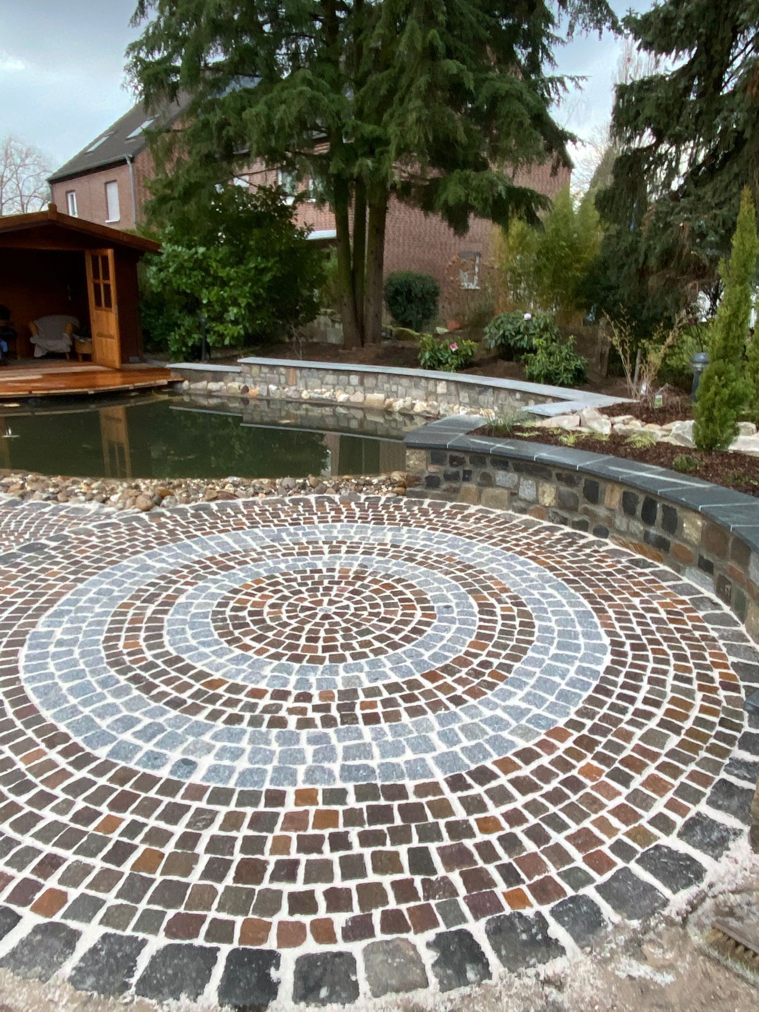 Ein runder, mit Kopfsteinpflaster belegter Patio liegt an einem Teich und ist von einer Steinmauer umgeben. Im Hintergrund befinden sich ein hölzerner Pavillon und Bäume.