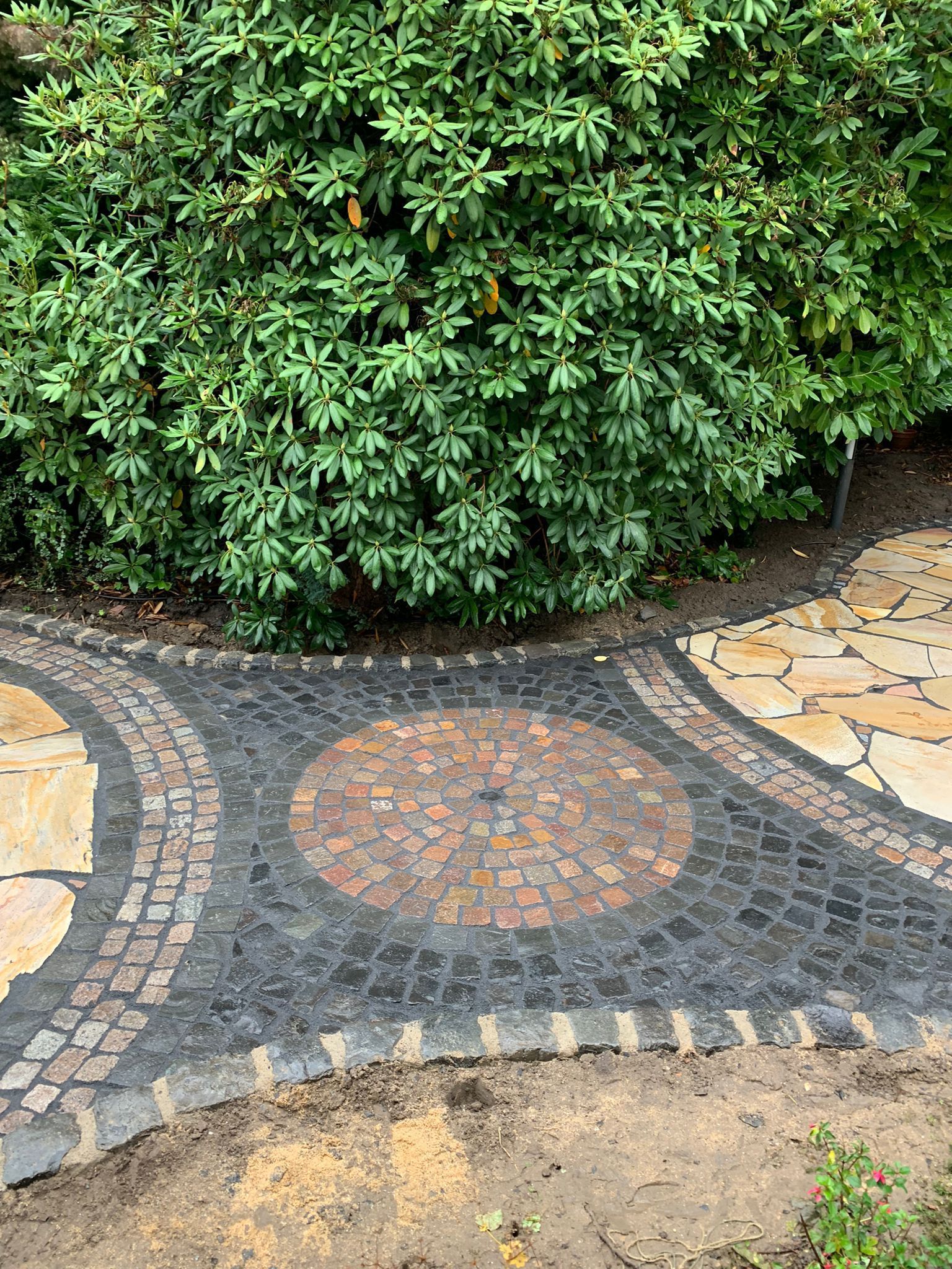 Gepflasterter Weg mit dekorativen schwarzen und braunen Steinen, der sich vor einem grünen Busch entlangschlängelt.