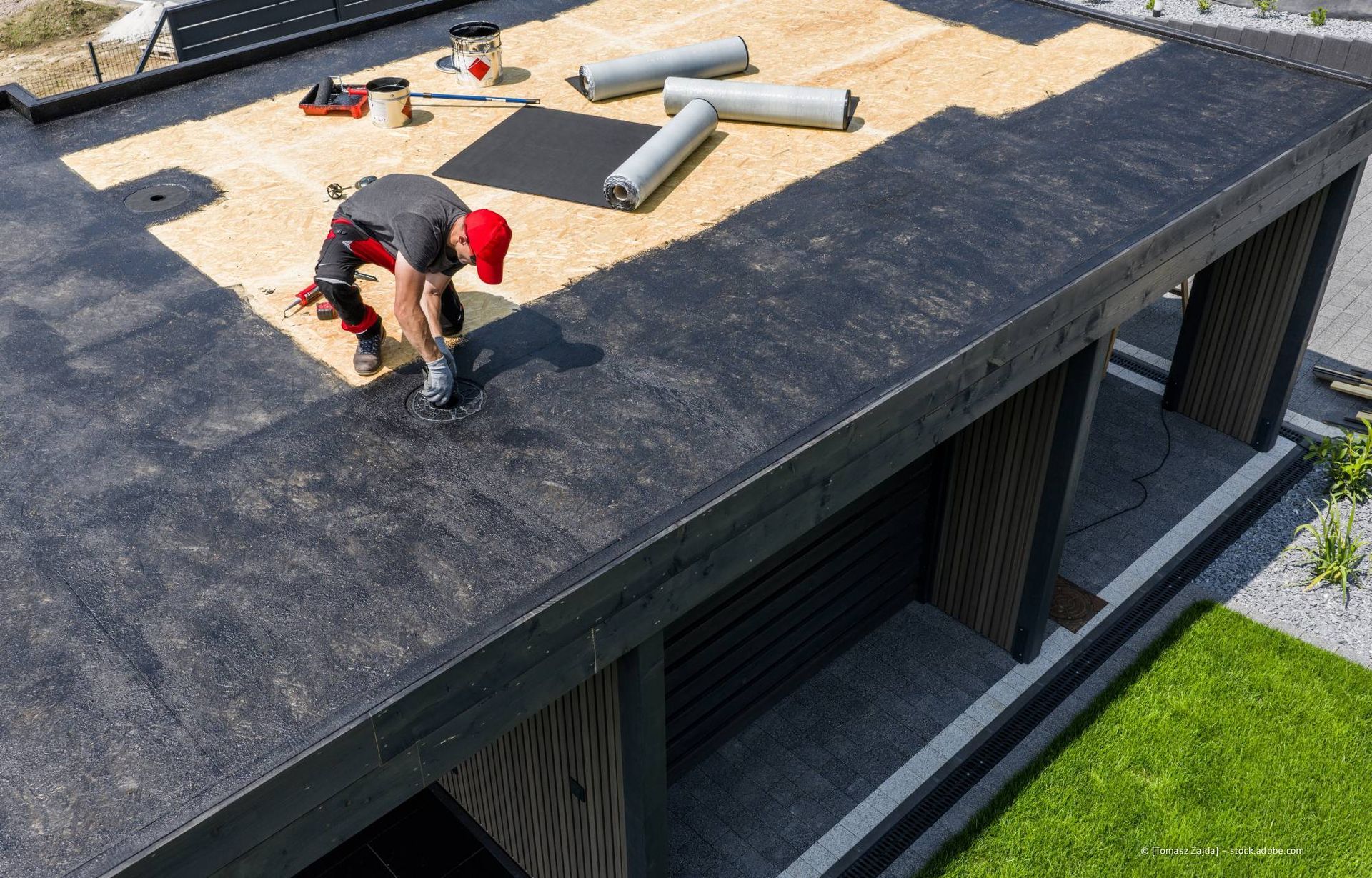 Ein Mann verlegt Dachmaterial auf einem schwarzen Flachdach. Materialrollen und Werkzeuge sind zu sehen.