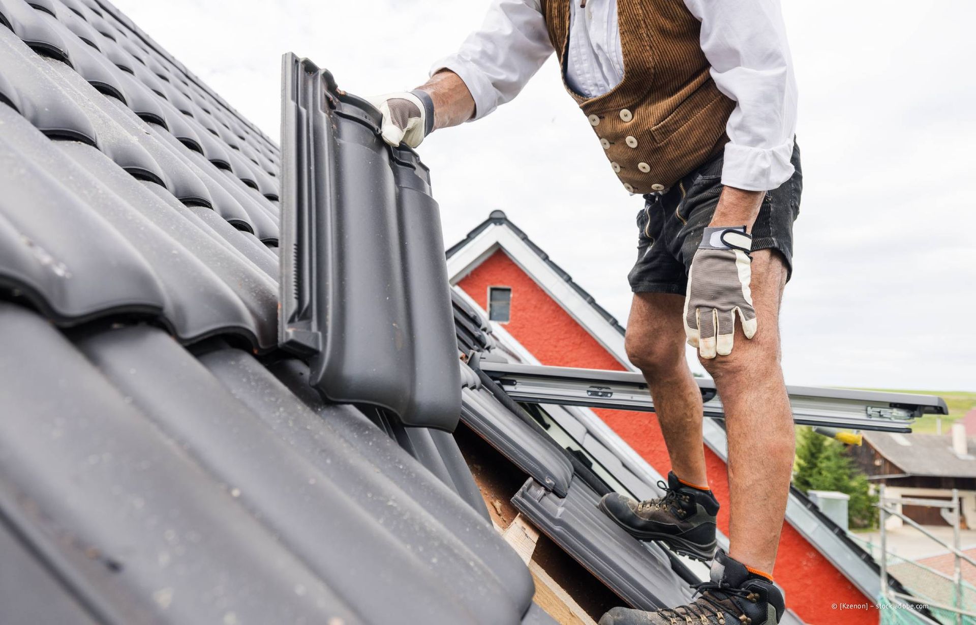 Ein Dachdecker verlegt dunkelgraue Dachziegel auf einem Schrägdach. Die Person trägt Schutzausrüstung und befindet sich im Freien.