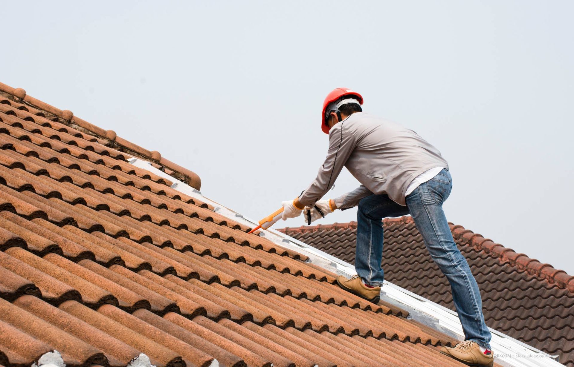 Ein Dachdecker verlegt dunkelgraue Dachziegel auf einem Schrägdach. Die Person trägt Schutzausrüstung und befindet sich im Freien.
