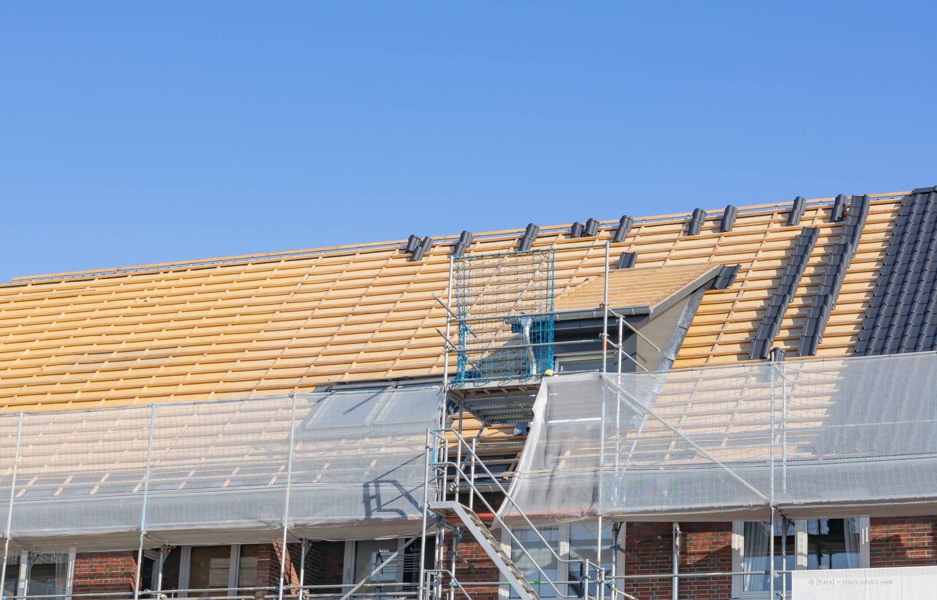 Ein Dachdecker verlegt dunkelgraue Dachziegel auf einem Schrägdach. Die Person trägt Schutzausrüstung und befindet sich im Freien.