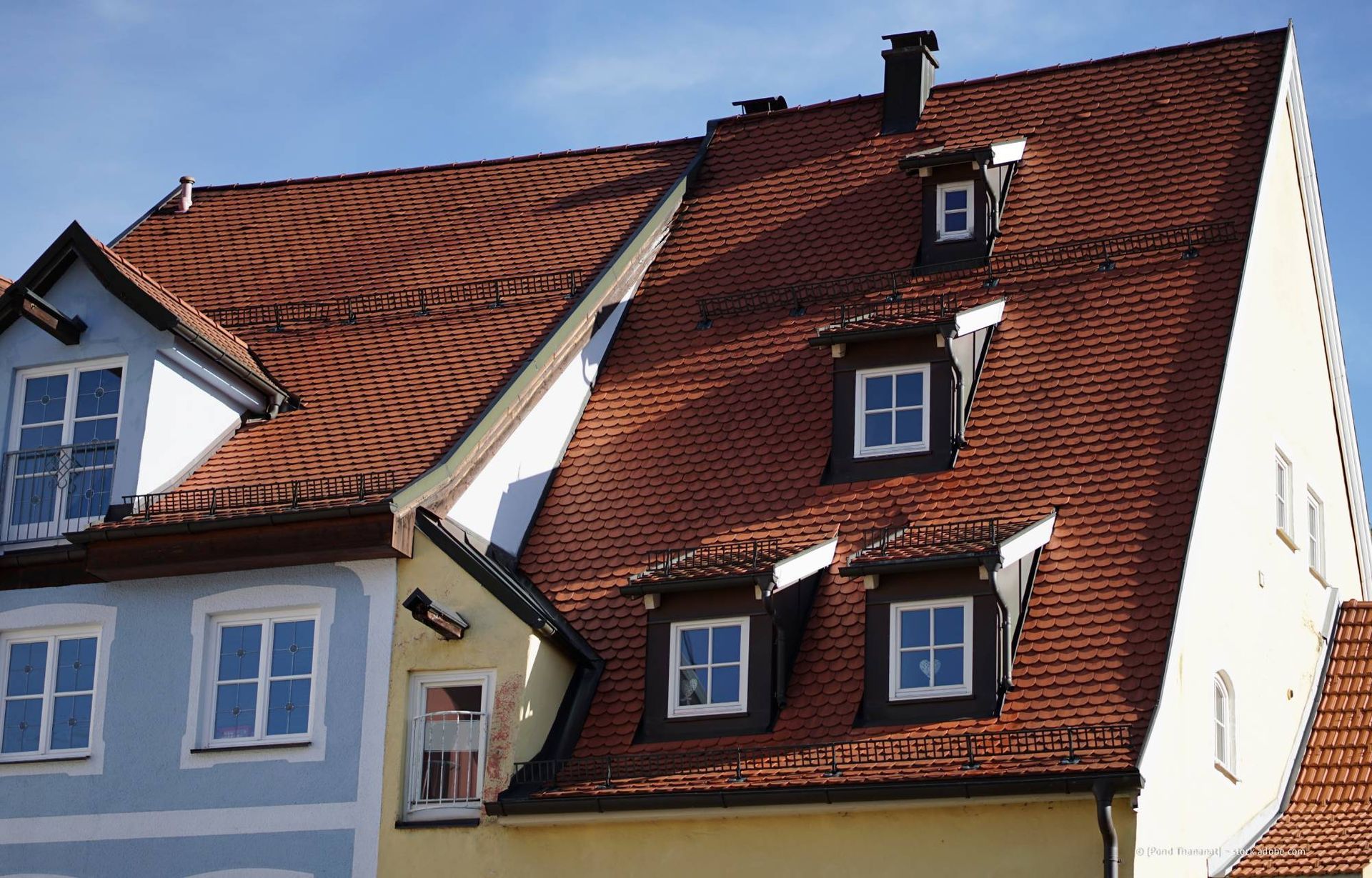 Gebäude mit roten Ziegeldächern und kleinen Dachgauben, blaue und gelbe Fassaden vor blauem Himmel.