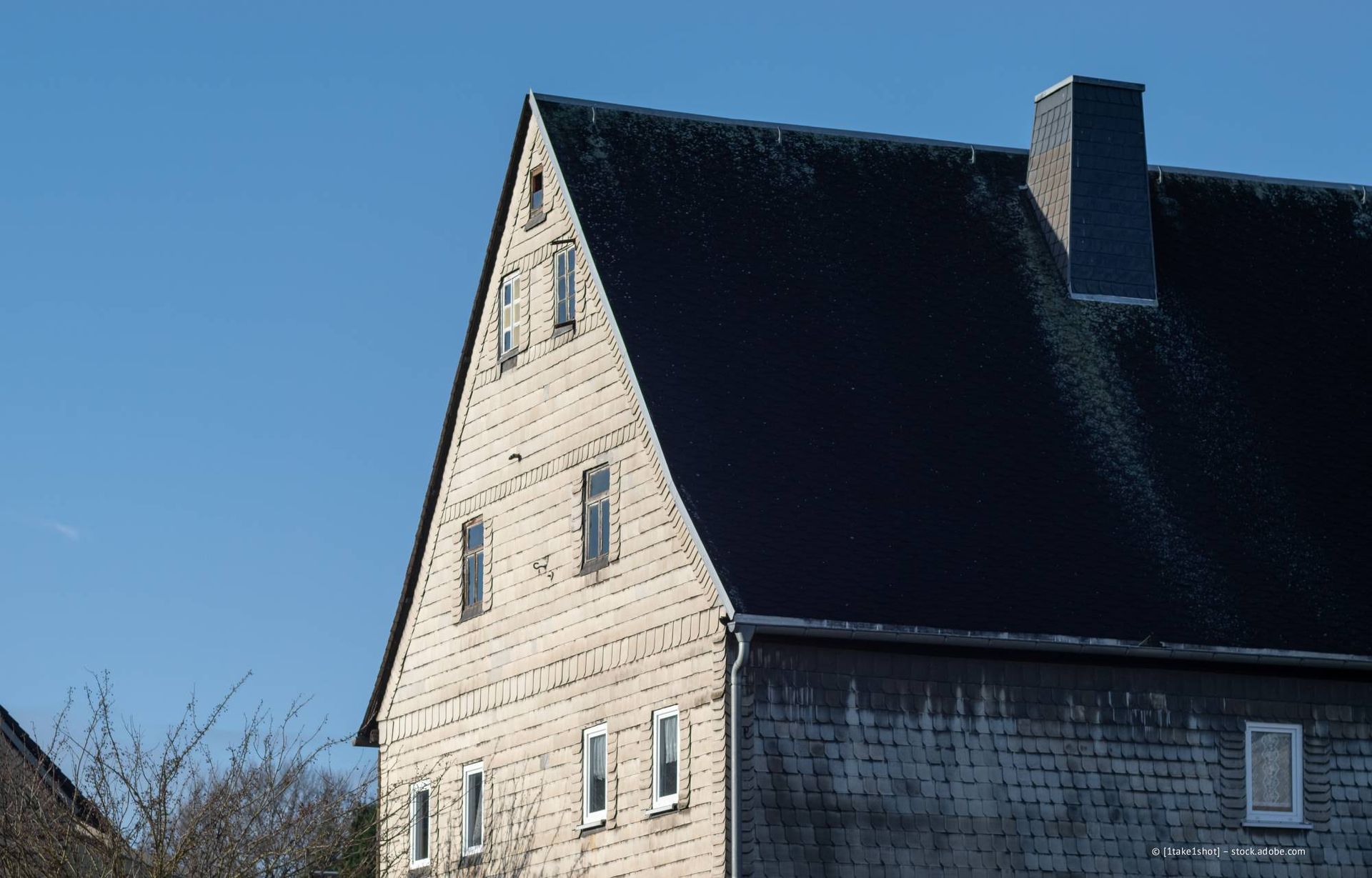 Ein Dachdecker verlegt dunkelgraue Dachziegel auf einem Schrägdach. Die Person trägt Schutzausrüstung und befindet sich im Freien.