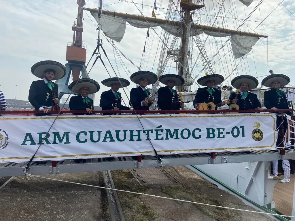 Tripulación con uniformes oscuros y sombreros a bordo de un barco, sosteniendo una pancarta que dice “ARM CUAUHTÉMOC BE-01”.