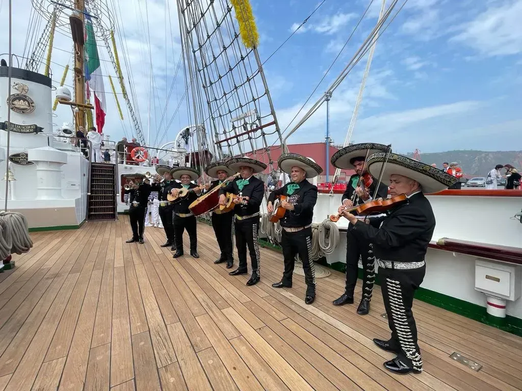 Un grupo de mariachis actuando en la cubierta de madera de un barco, bajo el aparejo y el cielo azul.