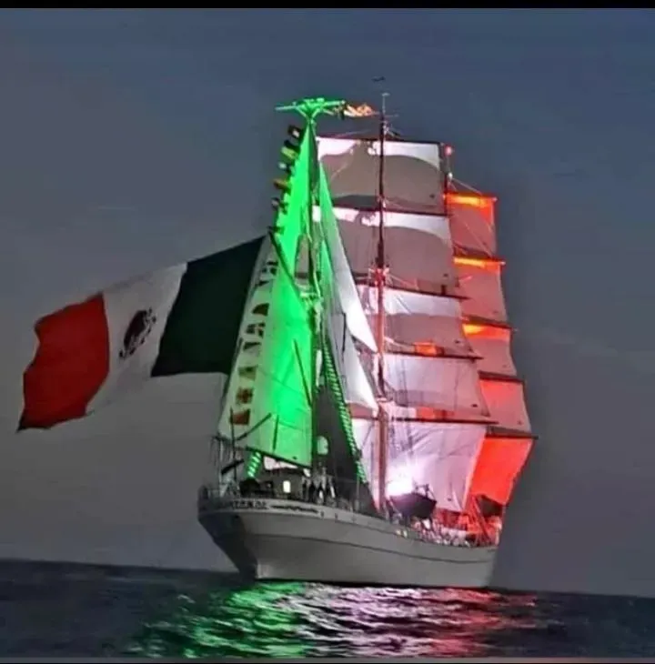 Un velero de noche iluminado en verde y rojo, con una gran bandera italiana ondeando sobre aguas oscuras.