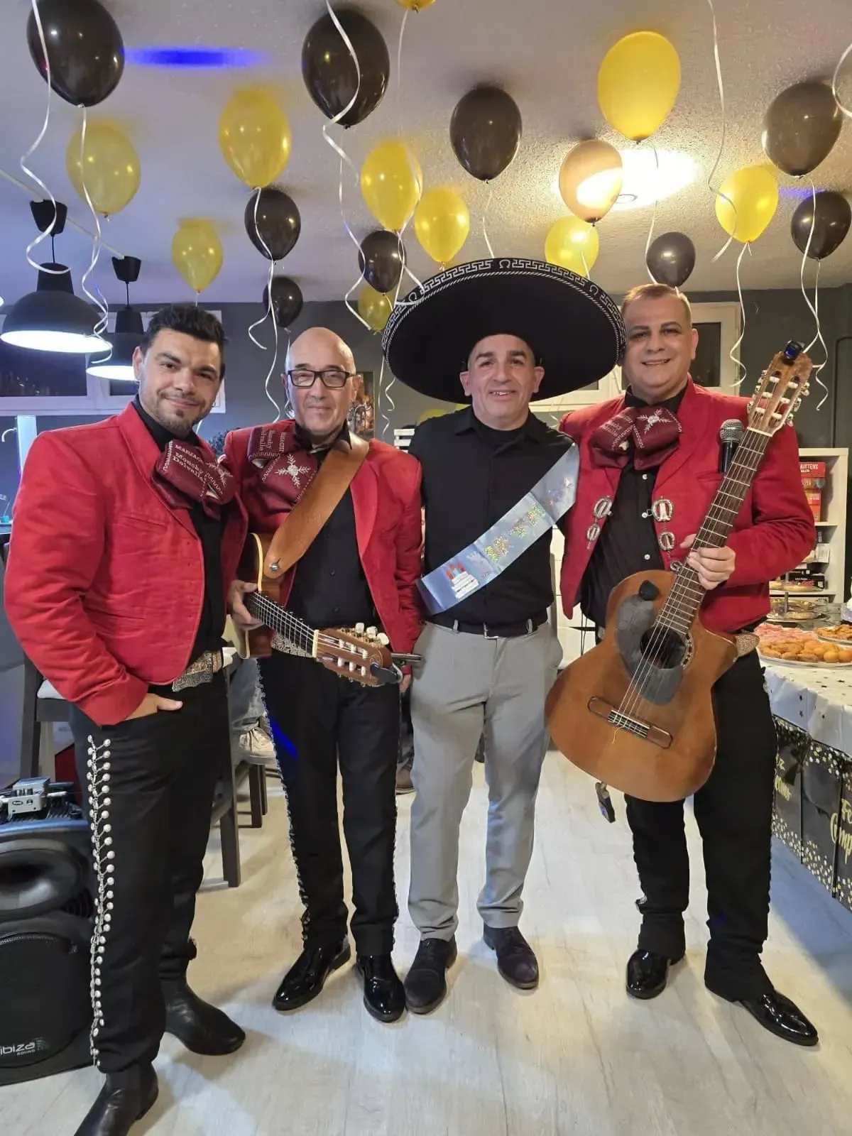 Cuatro músicos de mariachi con chaquetas rojas posan en el interior de un edificio, bajo globos negros, amarillos y blancos.
