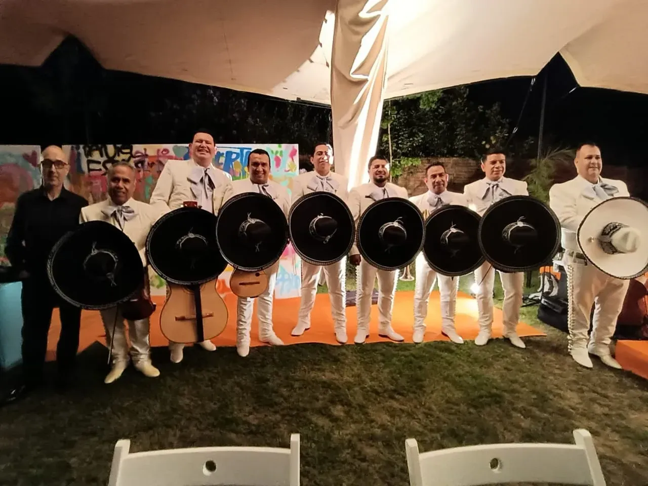 Grupo de personas vestidas de blanco sosteniendo grandes tambores negros bajo una tienda de campaña por la noche.