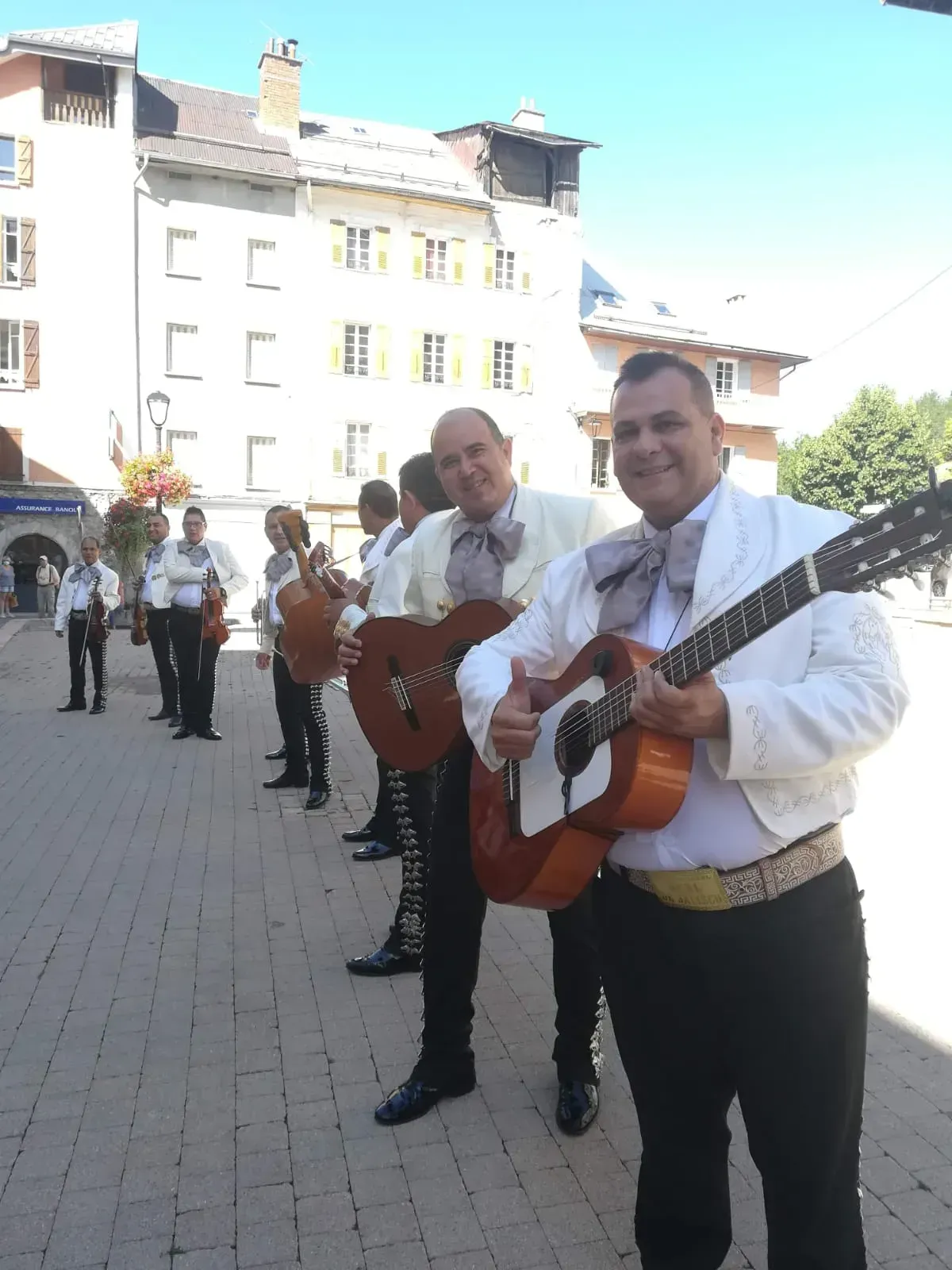 Músicos con camisas blancas tocando guitarras en una plaza soleada con edificios detrás.
