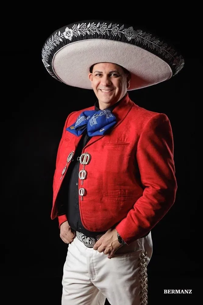 Hombre con traje de mariachi rojo y sombrero sonriendo sobre un fondo negro.
