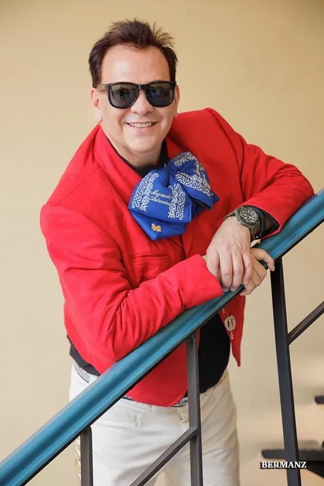 Hombre con chaqueta roja y bufanda azul apoyado en la barandilla de una escalera, con gafas de sol y sonriendo.
