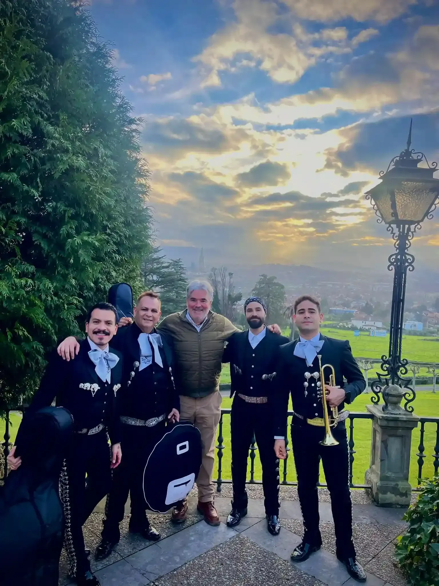 Cinco músicos con uniformes oscuros posan al aire libre al atardecer, junto a un sendero y una farola.