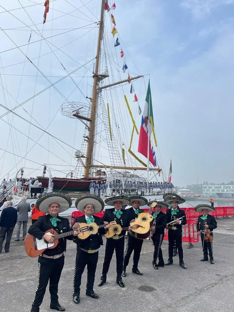 Un grupo de mariachis vestidos de traje negro toca la guitarra junto a un gran velero