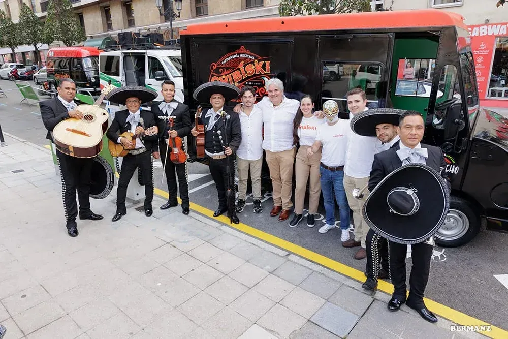 Músicos posando frente a un autobús rojo en una calle de la ciudad, sosteniendo guitarras, tambores y un sombrero.
