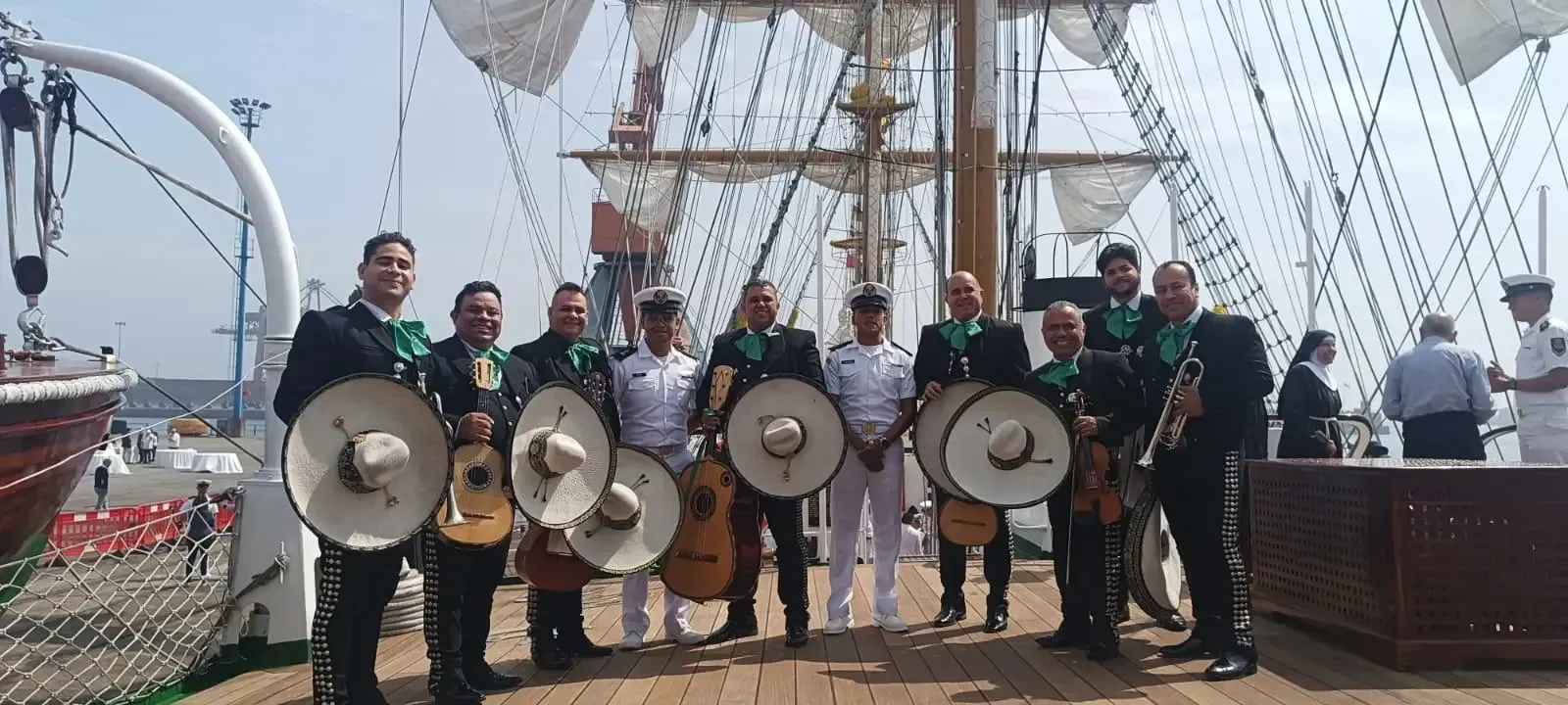 Grupo de personas sosteniendo escudos y posando en la cubierta de un barco con altos mástiles y aparejos al fondo.