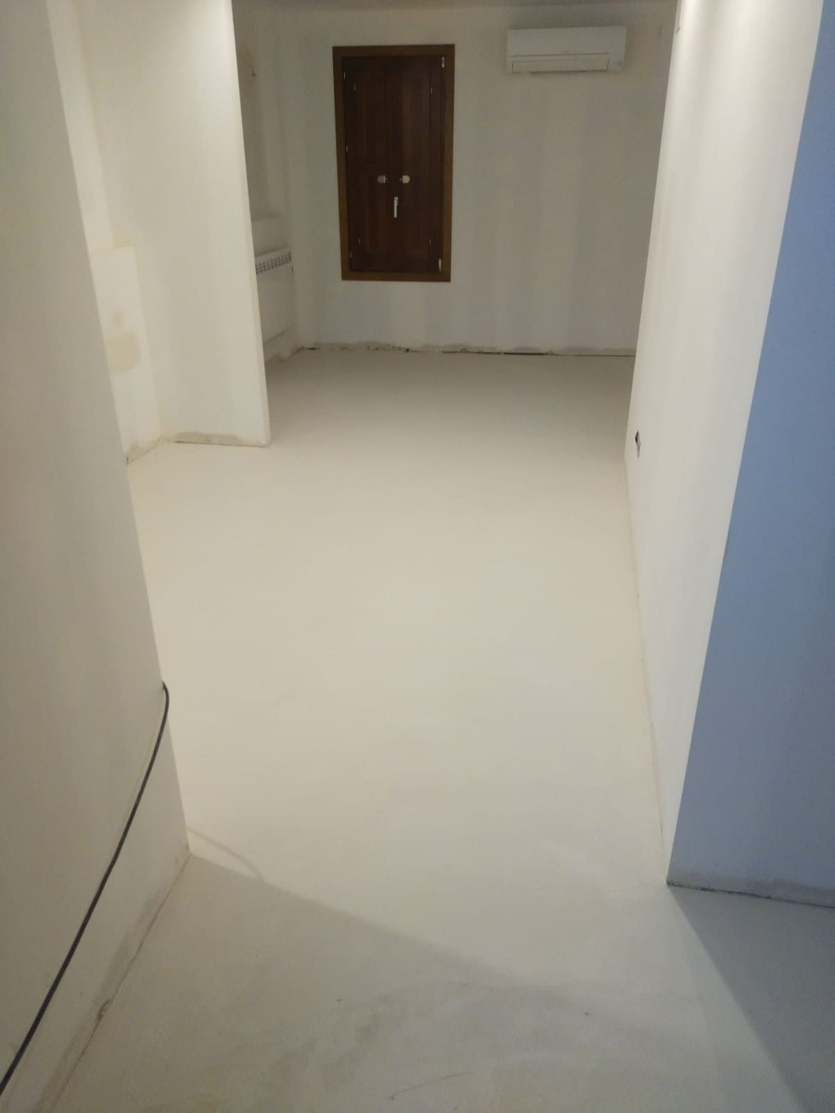 Pasillo vacío con suelo y paredes blancas, una puerta marrón al final y un marco de puerta azul a la derecha.