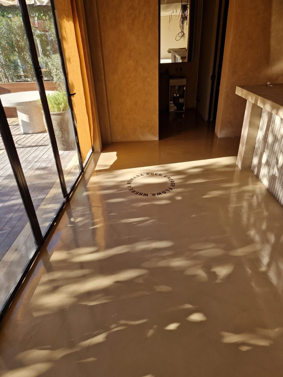 Pasillo interior iluminado por el sol con puerta de cristal abierta, suelo beige y sombras con patrones en el suelo.