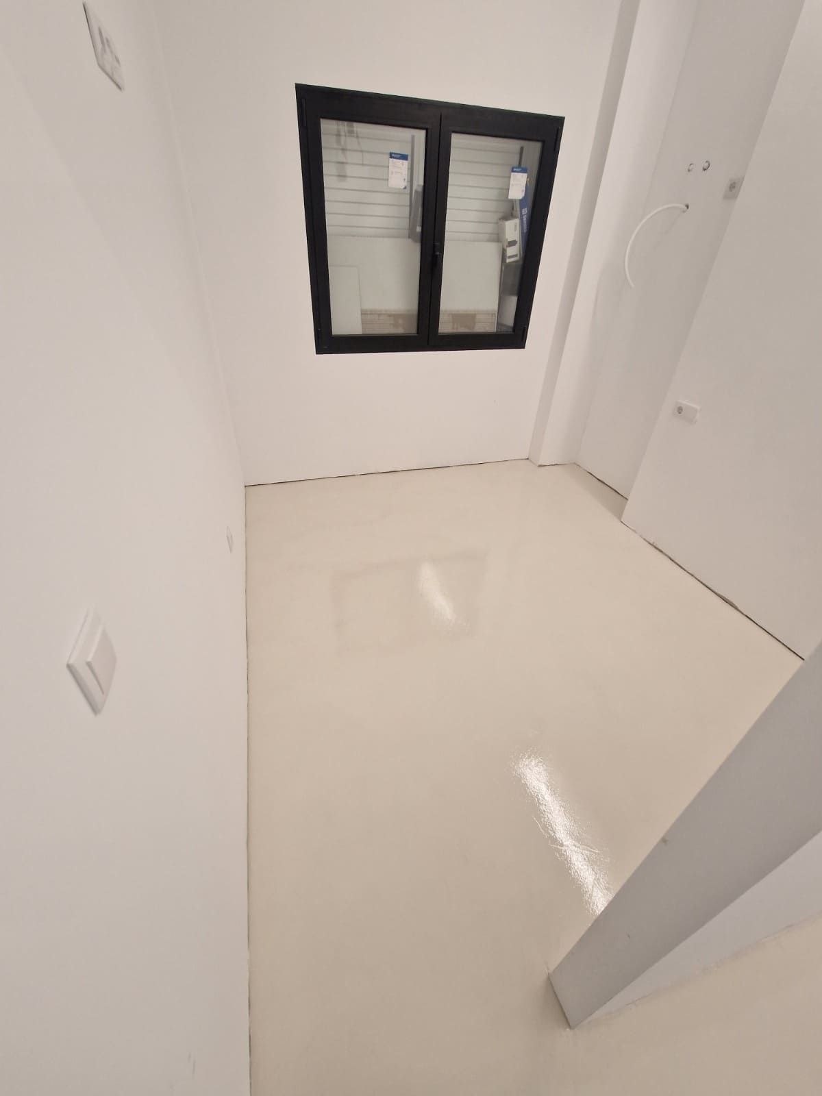 Habitación blanca vacía con suelo brillante, ventana con marco negro y puertas blancas cerradas.