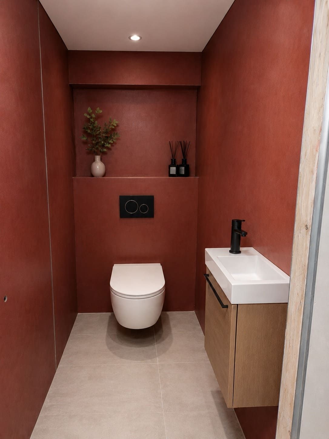 Baño moderno y estrecho con paredes rojas, inodoro blanco y lavabo pequeño con mueble de madera.