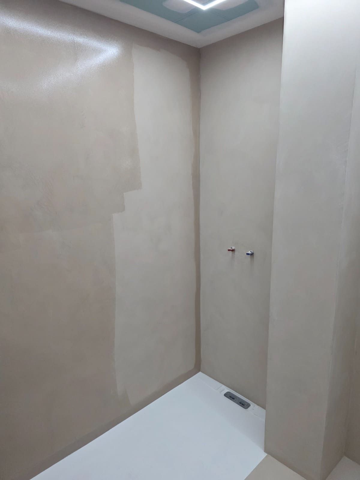 Cabina de ducha vacía de color beige con suelo blanco y dos manijas montadas en la pared cerca de un desagüe.