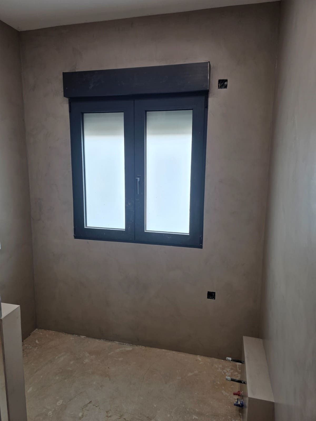Habitación vacía sin terminar con paredes grises, una ventana con marco azul y suelo desnudo en construcción.