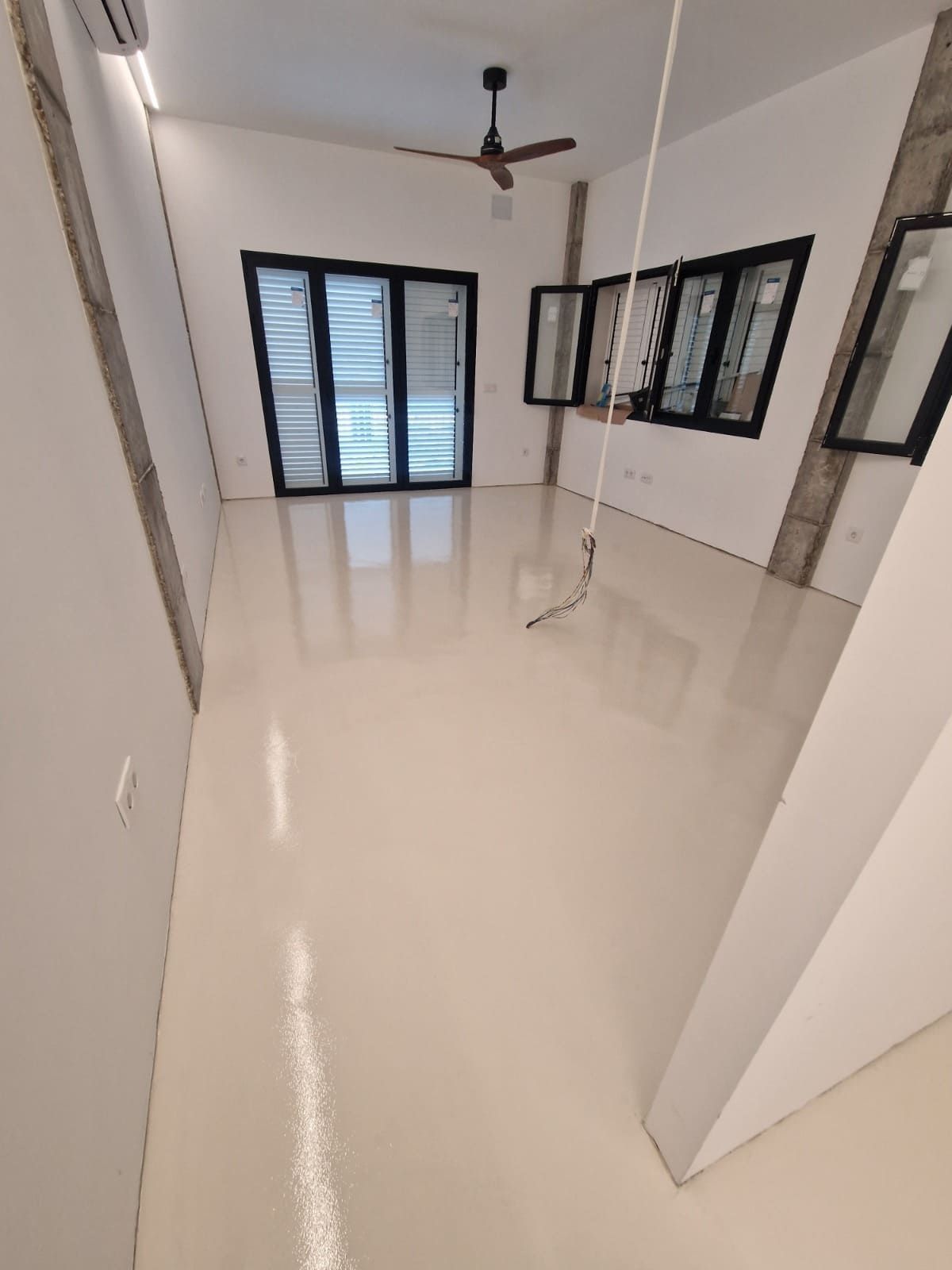 Habitación luminosa y vacía con suelo brillante, ventilador de techo y grandes ventanas y puertas con marcos negros.