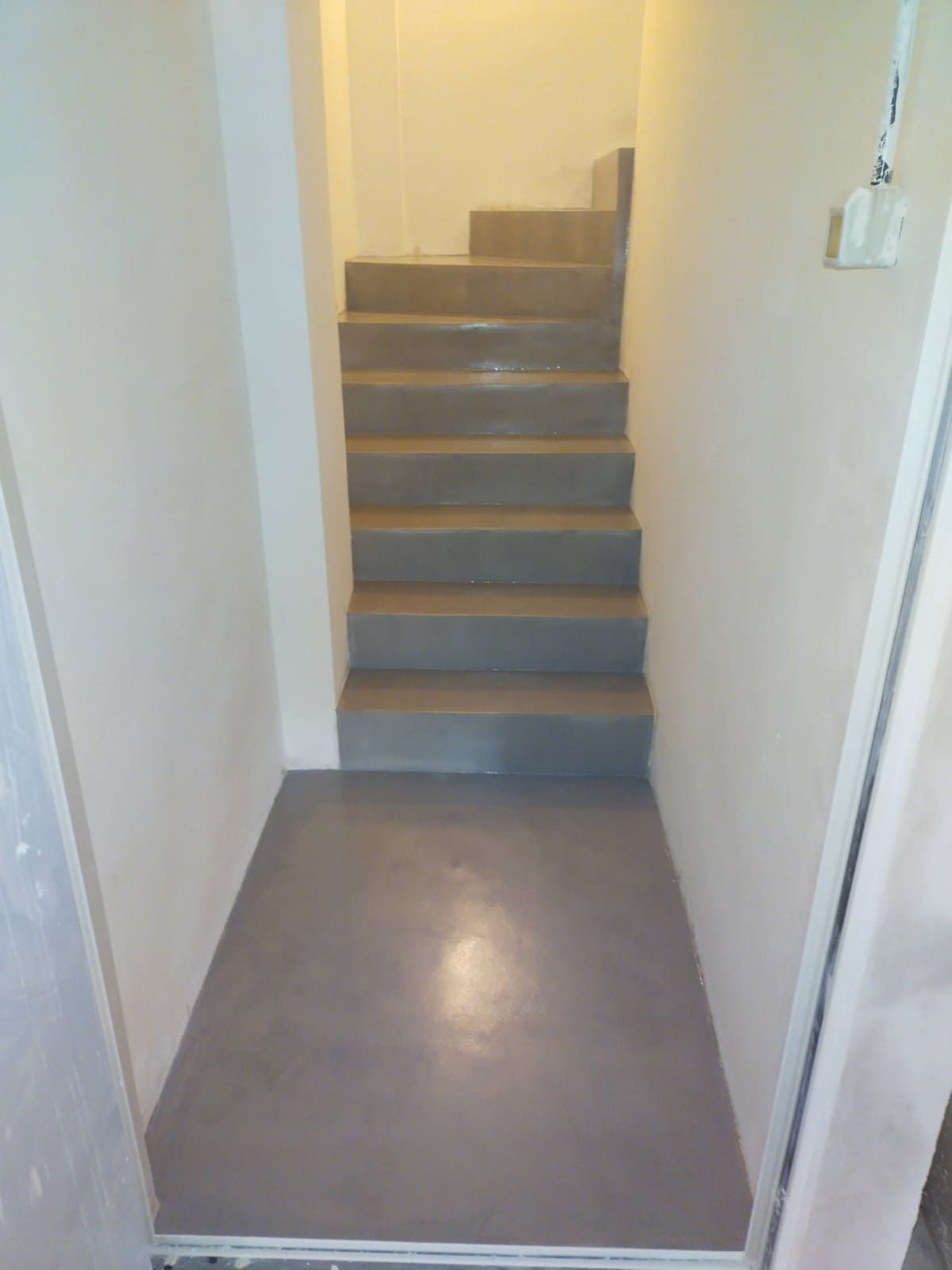 Estrecha escalera interior con escalones grises que asciende desde un pequeño pasillo.