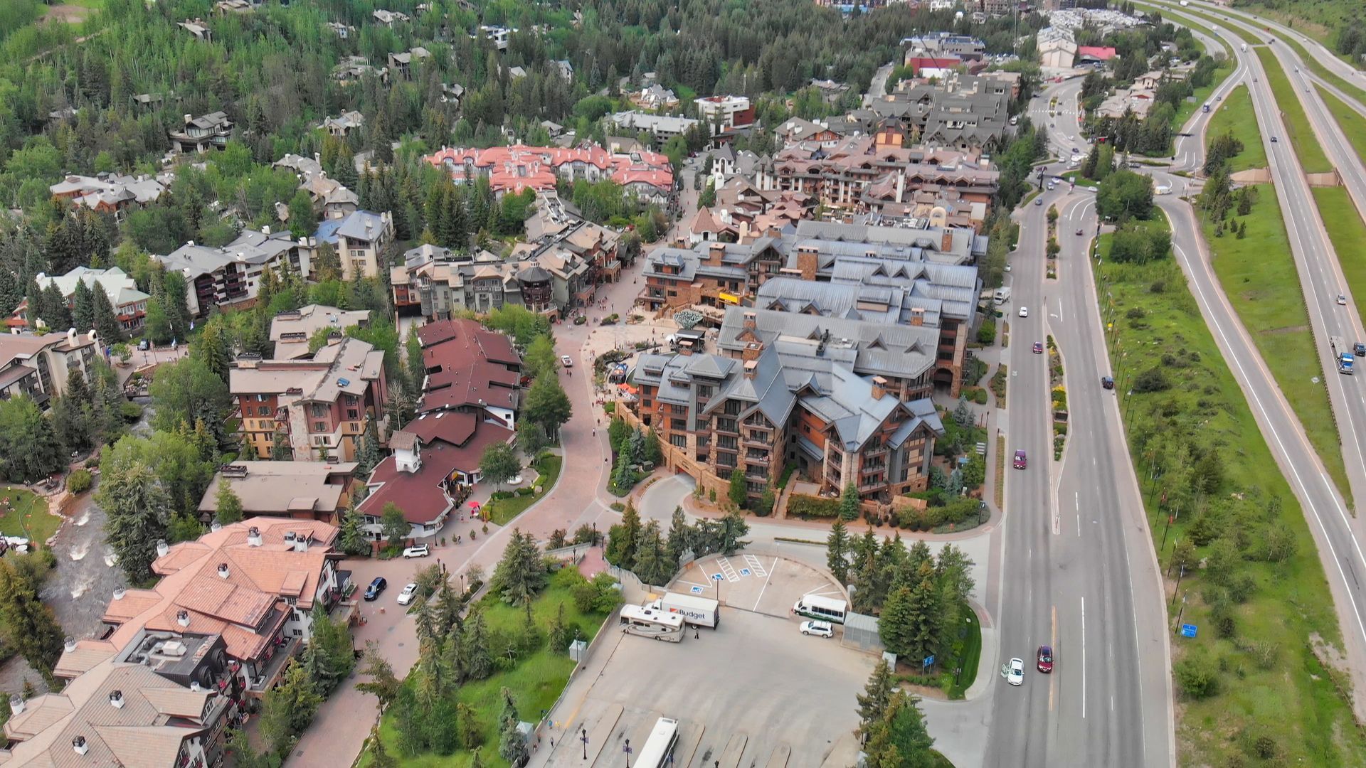 Aerial view of a mountain resort with buildings, parking lot, and a highway beside forested hills.