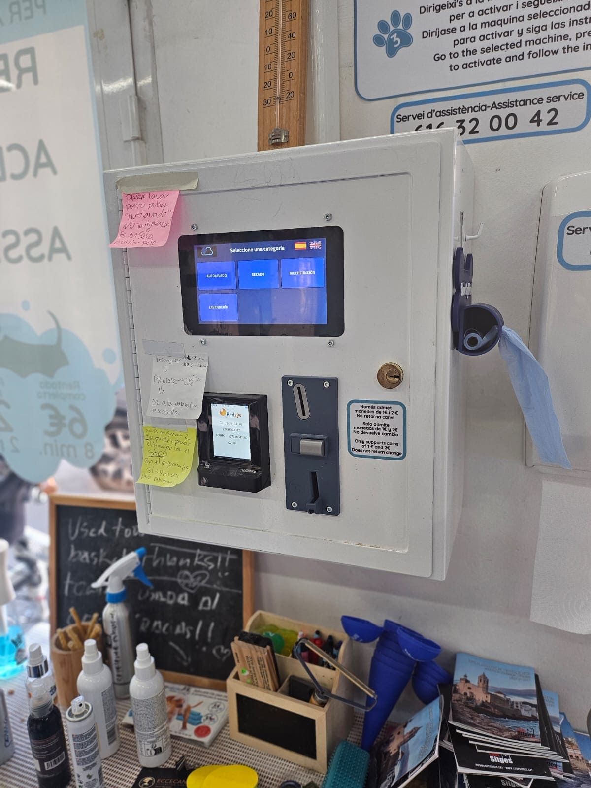 Un quiosco de pago para máquinas expendedoras, de color blanco, montado en la pared, con pantalla táctil, lector de tarjetas y ranura para monedas, ubicado en una tienda de servicios.