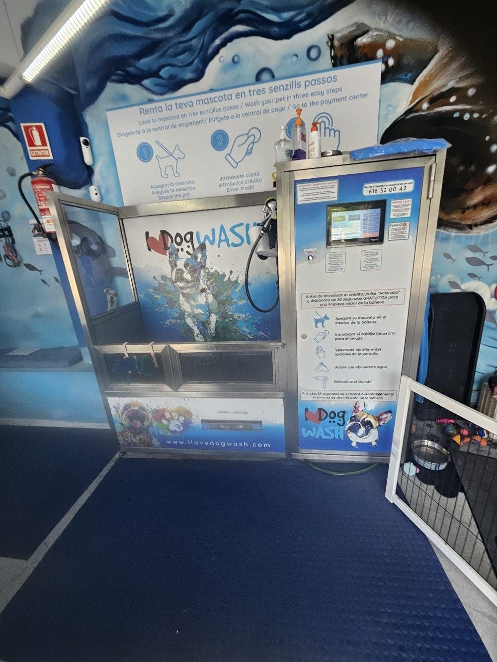 Una estación de lavado de perros de autoservicio en interiores con una tina de metal, un panel de control con pantalla y señalización instructiva en la pared.