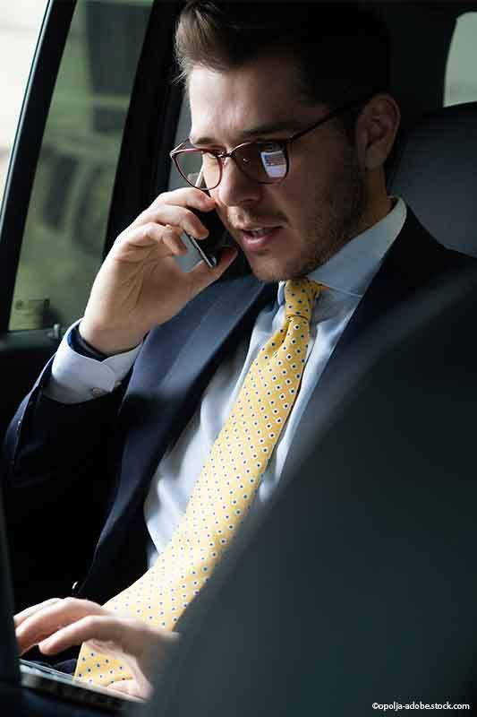 Mann im Anzug, Brille, telefoniert im Auto, arbeitet am Laptop. Gelbe Krawatte mit schwarzen Punkten.