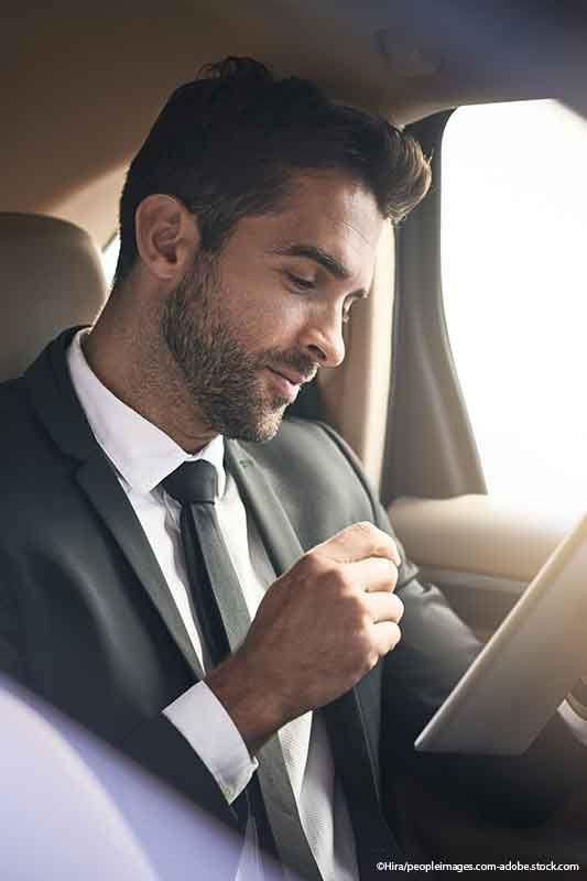 Mann im Anzug in einem Auto, der mit nachdenklichem Gesichtsausdruck auf ein Tablet blickt.