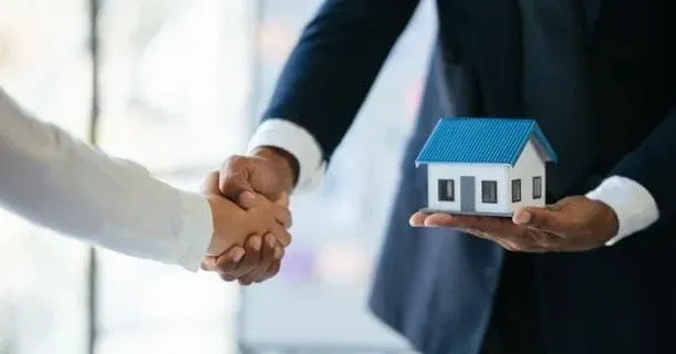 Duas pessoas apertando as mãos, uma delas segurando uma casa em miniatura; provavelmente uma transação imobiliária.