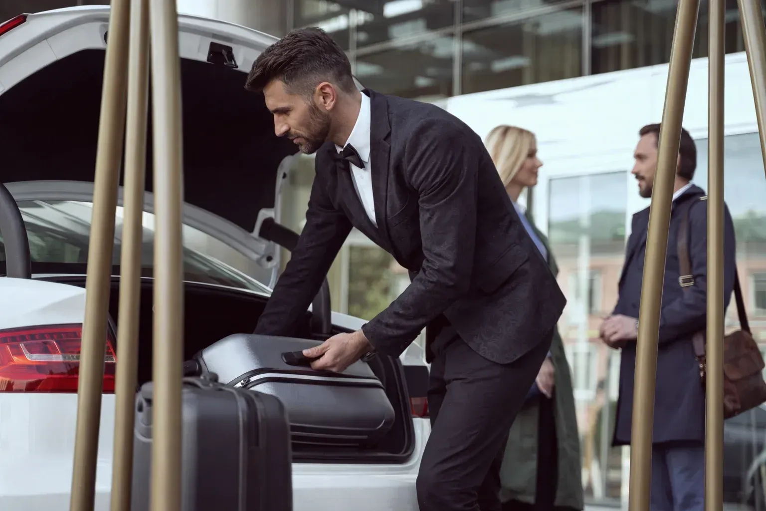 A person in a suit loads luggage into the trunk of a car while two people stand nearby outside a modern building.