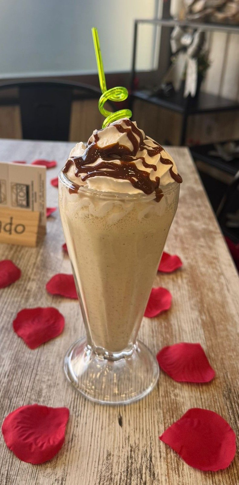 Batido de chocolate con sirope en un vaso alto, coronado con nata montada y una pajita verde, sobre una mesa.