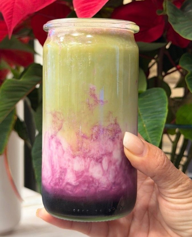 Una mano sostiene un tarro de cristal con una bebida o postre en capas de color morado, rosa y verde pálido.