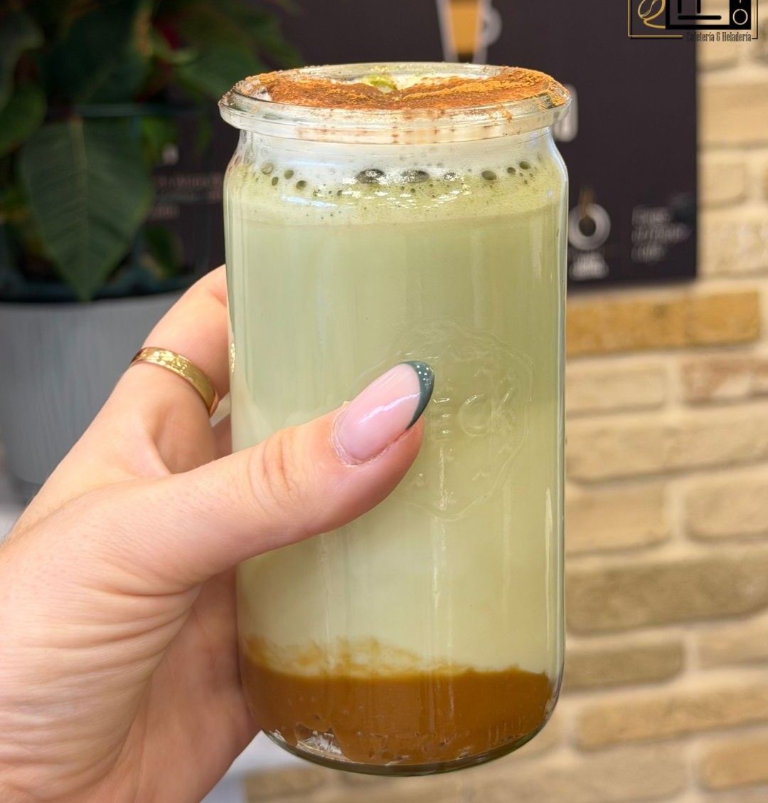 Una mano sostiene una bebida helada espumosa en capas dentro de un frasco de vidrio transparente, con jarabe marrón de fondo.
