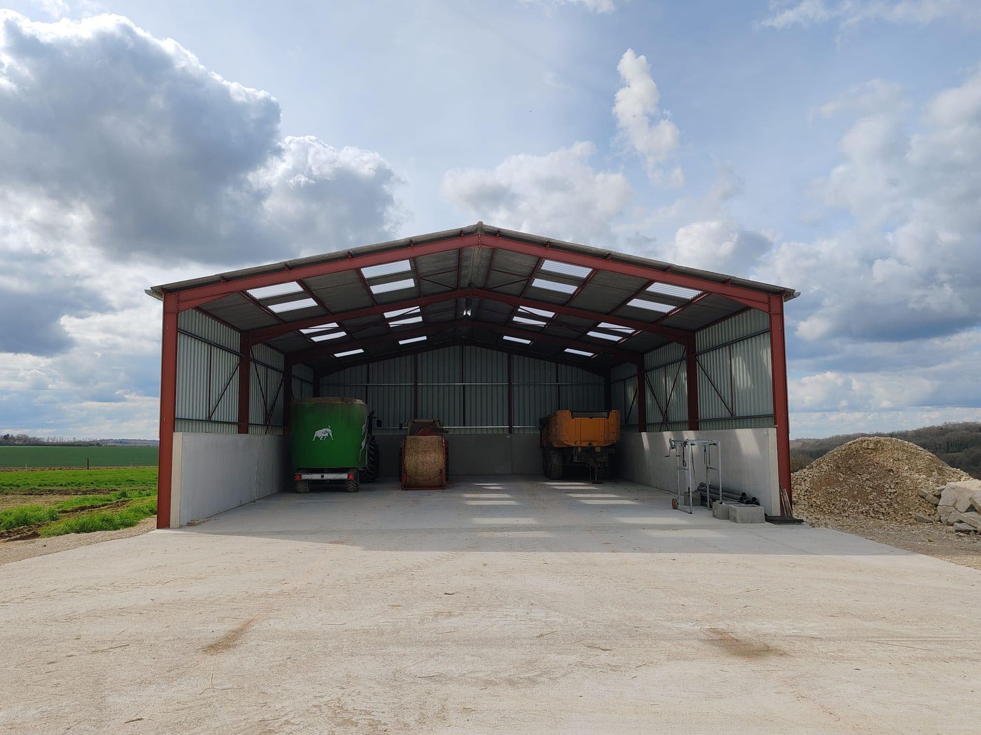 Hangar agricole ouvert sur les côtés, avec un toit en métal, des bacs et des tonneaux à l’intérieur, dans une cour gravillonnée sous un ciel nuageux
