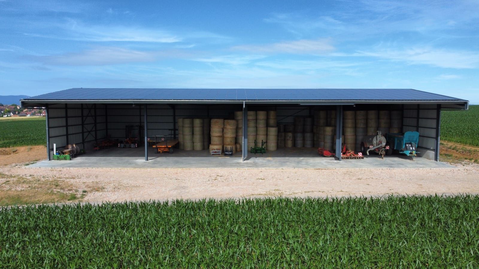 Long hangar bas et allongé, semblable à une grange, avec un toit bleu et une façade ouverte, au bord d’un champ verdoyant.