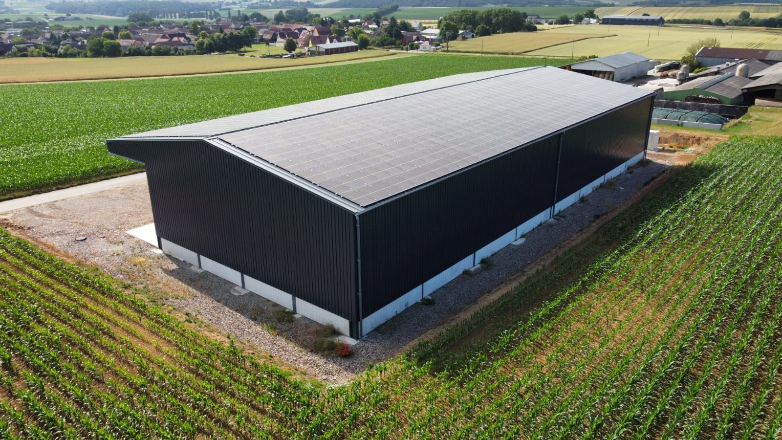 Vue aérienne d’un long bâtiment agricole noir au toit gris, bordé de champs verdoyants