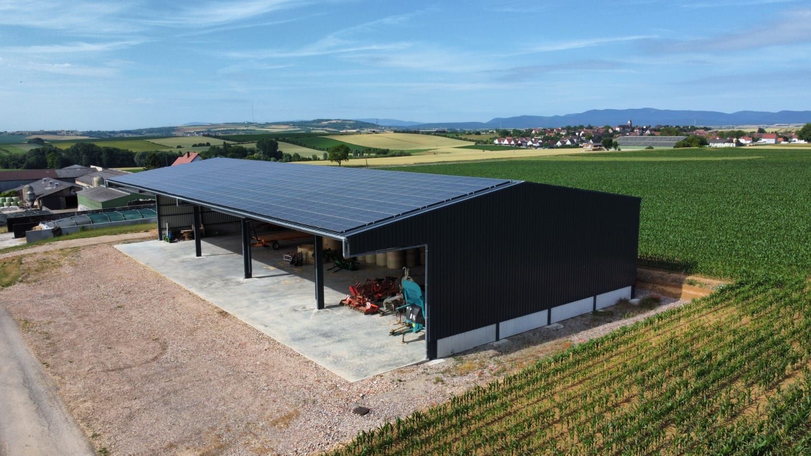 Hangar agricole noir avec un toit solaire, au bord de champs cultivés sous un ciel dégagé
